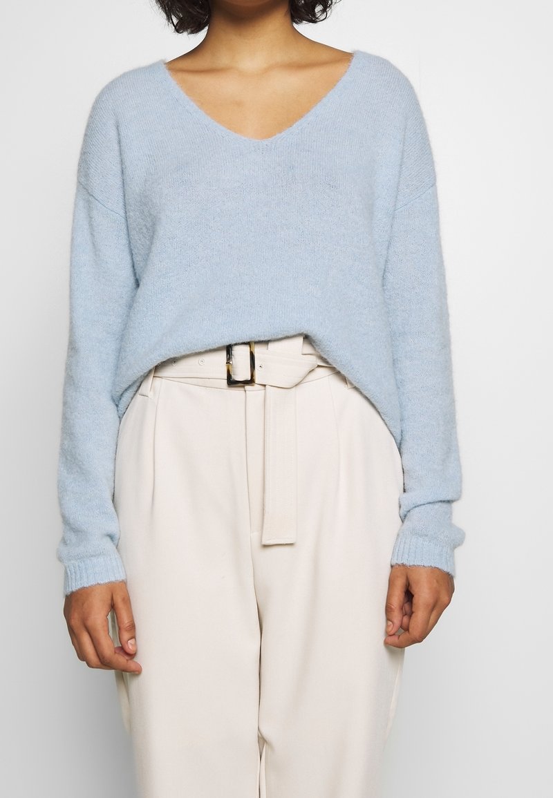 Jasnoniebieski sweter z dekoltem w serek o luźnym kroju i ściągaczami na rękawach, zestawiony z kremowymi, wysokim stanem spodniami i pasującym paskiem.