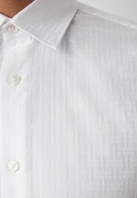 Weißes Hemd mit strukturiertem Design und subtilen geometrischen Mustern, Knopfkragen und einem einzelnen Knopf vorne.