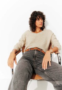 Pull en tricot beige avec un col en V, jean gris taille haute, ceinture en cuir marron, et une montre métallique, assis sur une chaise en chrome.