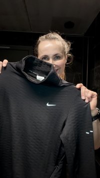 Haut Nike noir à manches longues avec un col montant, des rayures texturées, un logo réfléchissant et des accents latéraux. Présenté dans un environnement intérieur peu éclairé.