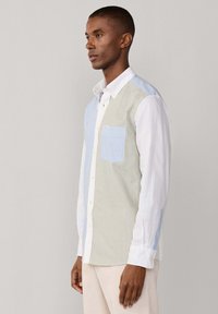 Uomo che indossa una camicia a maniche lunghe con bottoni e strisce verticali miste blu, verdi e bianche e pantaloni beige, in piedi di lato.