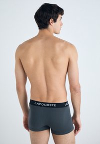 Graue Boxershorts mit einem schwarzen elastischen Bund, auf dem "LACOSTE" in weißer Schrift steht, mit glatter Textur und enganliegendem Design.