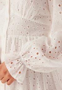 Weißes Kleid aus Lochstickerei mit kleinen runden Ausschnitten, mit Puffärmeln und elastischen Bündchen sowie einer gerafften Taille.