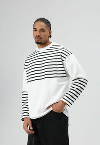 Witte sweatshirt met horizontale zwarte strepen over het bovenste deel en de mouwen. Zachte textuur, losse pasvorm, gecombineerd met zwarte broek.
