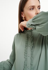 Blusa verde claro con un placket abotonado con volantes, tejido suave y mangas abullonadas. Detalle de encaje notable a lo largo del frente.