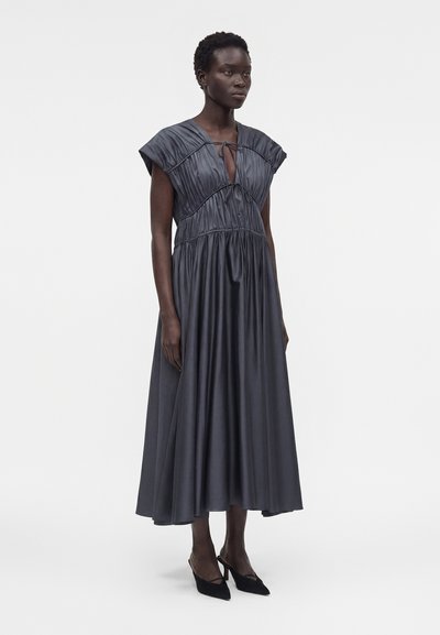 Γυναίκα που φοράει ένα σκούρο γκρι, πλισέ midi φόρεμα με μανίκια cap και μπροστινό δέσιμο σε V λαιμόκοψη, σε συνδυασμό με μαύρες μυτερές γόβες.