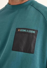 Sudadera verde azulado con un bolsillo negro que presenta el texto "CONVIÉRTETE EN UNA LEYENDA" en turquesa y naranja. Material suave de mezcla de algodón, cuello acanalado.