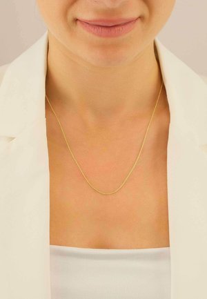 Collana a catena d'oro con un design delicato e intrecciato, posata su una clavicola scoperta, abbinata a un blazer bianco e a una maglietta.