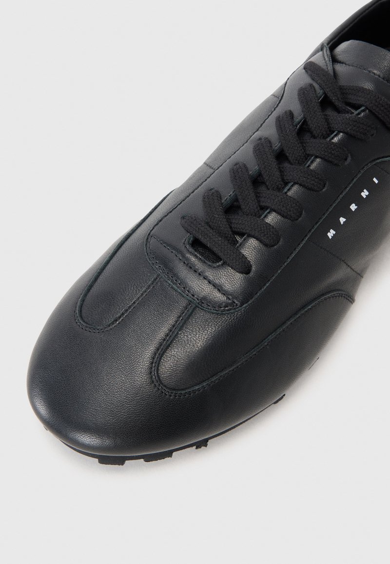 Schwarze Leder-Sneaker mit glatter Oberfläche, mit abgerundetem Zehenbereich, einfarbiger Nähte und flachen schwarzen Schnürdetails. Markennamen seitlich aufgedruckt.
