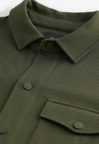 Camisa verde texturizada con cuello de solapa, botones a presión y un bolsillo en el pecho con un acento oscuro.