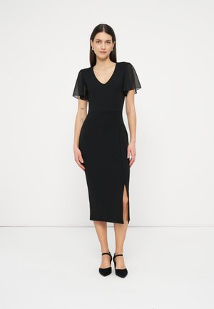 Žena v čiernych midi šatách s priesvitnými plápolajúcimi rukávmi, výstrihom do V, postranným rázporkom a čiernymi lodičkami s remienkom okolo členka, stojaca pred jednoduchým bielym pozadím.