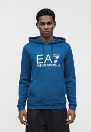 Jeune homme portant un sweat à capuche bleu EA7 Emporio Armani avec une poche avant, debout devant un fond clair uni.