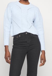 Maglione lavorato a maglia di colore blu chiaro con texture a coste e dettagli in pizzo, abbinato a jeans neri aderenti con tasche classiche e chiusura a bottone.