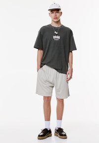 Ung mand iført en hvid kasket, en mørk oversized Honda T-shirt, lyse løse shorts, hvide sokker og sorte klodsede sko, står op ad en hvid baggrund.
