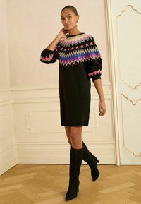 Rochie tricotată neagră cu un model geometric colorat la decolteu, cu sleeves voluminoase și asortată cu cizme negre până la genunchi.