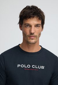 Mörkblå bomulls-t-shirt med kort ärm och rund halsringning. Har vitt och rött broderat text: "POLO CLUB ESTABLISHED 2012." Mjuk textur.