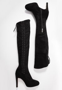 Bottes en suédine noires au-dessus du genou avec un talon aiguille. Comprend une fermeture éclair sur un côté et un détail de lacet à l'arrière pour un ajustement réglable.