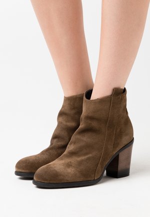 Stivali alla caviglia in suede marrone con tacco a blocco indossati su gambe nude su uno sfondo bianco.