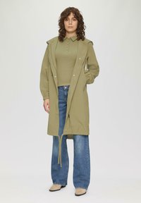 Khaki lange jas met een capuchon, twee zijzakken en een taille met trekkoord, gelaagd over een groene top en uitlopende spijkerbroek.
