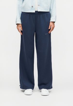 Personne portant un pantalon de survêtement large bleu marine, des baskets blanches et une veste en jean bleu clair, debout devant un fond uni.