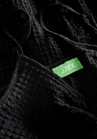 Tessuto nero con texture e motivo a increspature. Presenta spalline sottili e un'etichetta verde del marchio "JJXX."