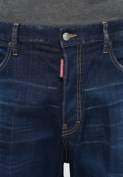 Jean en denim bleu foncé avec un motif de décoloration distinct, des coutures jaunes et une étiquette de logo rouge sur la ceinture. Fermeture à boutons.