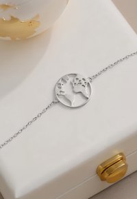 Silverarmband med en rund globhängsmycke med utklippta kontinenter. Kedjan är länkad och ligger på en beige låda med ett guldspänne.