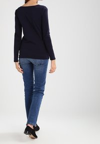 Marineblauw langdunne top met een bootnek, gecombineerd met aangesloten blauwe jeans en zwarte schoenen met een lage, glanzende hak. Effen stof, geen patronen.
