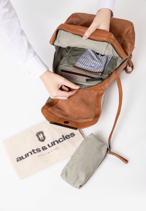 Bruine leren rugzak met een ritssluiting, gevoerd interieur van stof en zakken. Inclusief een beige canvas tas met logo en een kleine groene tas.