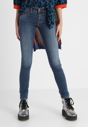 Femme portant un jean skinny bleu, un pull à imprimé léopard avec des poignets orange et des bottes à plateforme argentée à lacets, debout devant un fond blanc.