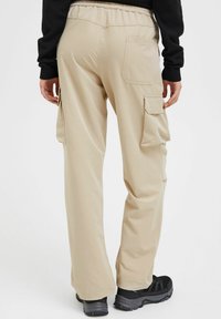 Pantalons cargo beiges avec une taille élastique, poches latérales et arrière, et une coupe décontractée. Portés avec des chaussures noires sur un fond clair.