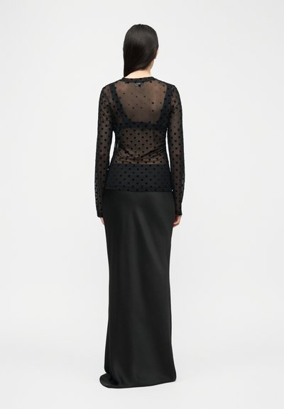 Γυναίκα με μακριά σκούρα μαλλιά, φοράει μαύρο διαφανές μακρυμάνικο τοπ με σχέδια καρδιών πάνω από μαύρο κορμάκι και μακριά μαύρη φούστα, γυρισμένη προς τα πίσω.