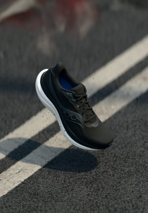 Zapatilla de running negra con suela blanca suspendida sobre una carretera asfaltada con marcas blancas de carril, con pequeños escombros dispersos debajo.