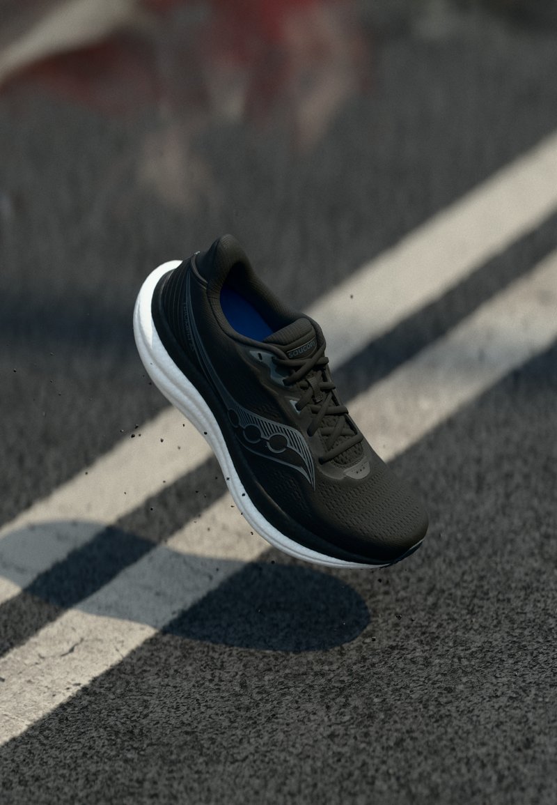 Μαύρο αθλητικό παπούτσι με λευκή σόλα να αιωρείται πάνω από δρόμο με άσφαλτο και λευκές διαγραμμίσεις, με μικρά υπολείμματα να σκορπίζουν από κάτω του.
