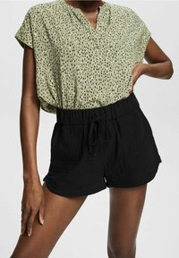 Žena na sobě má zelenou tečkovanou blůzu s krátkým rukávem, která je zastrčená do černých šortek na šňůrku. Stojí s jednou rukou na boku a druhou rukou uvolněnou.