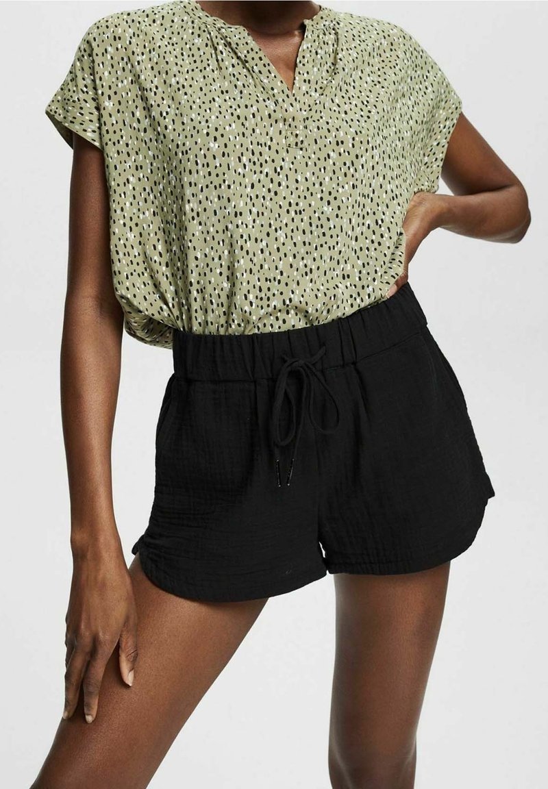 Žena na sobě má zelenou tečkovanou blůzu s krátkým rukávem, která je zastrčená do černých šortek na šňůrku. Stojí s jednou rukou na boku a druhou rukou uvolněnou.