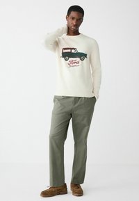 Kreemvärvi sviiter, millel on Ford Bronco graafika tumerohelises ja punases. Koos oliivroheliste pükste ja pruunide mokassini-stiilis kingadega.