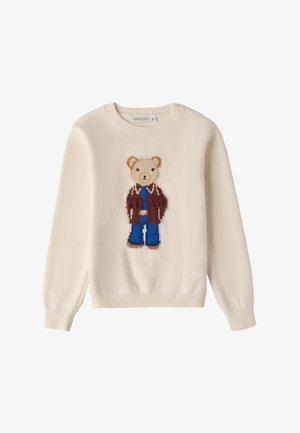 Pull crème orné d'un motif d'ours en peluche vêtu d'une veste brune et d'un pantalon bleu, avec une texture tricotée et des poignets côtelés.