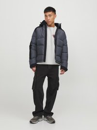 Giubbotto imbottito grigio scuro con cappuccio, dettagli neri, design trapuntato e zip frontale. Abbinato a pantaloni cargo neri larghi e sneakers.