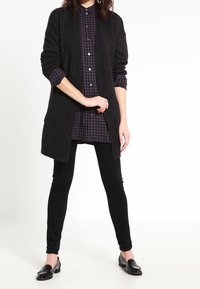 Cardigan lungo nero sopra una camicia a quadri scuri, abbinato a jeans skinny neri e mocassini neri. Texture morbida e vestibilità rilassata.