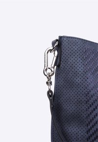 Coin d'un sac en tissu perforé bleu avec un mousqueton en métal argenté et une sangle noire attachée, sur fond blanc.