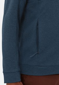 Giacca in pile blu con texture, caratterizzata da una tasca laterale con zip, polsini a costine e un orlo con coulisse, progettata per calore e comfort.