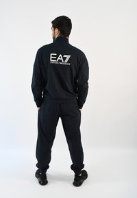 Chándal azul marino que incluye una chaqueta con cremallera y pantalones ajustados, con un logo blanco de EA7 en la parte posterior. El material parece suave y casual.