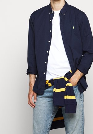 Mann trägt ein aufgeknöpftes marineblaues Hemd über einem weißen T-Shirt, hellblaue Jeans und einen marineblau-gelb gestreiften Pullover, um die Taille gebunden.