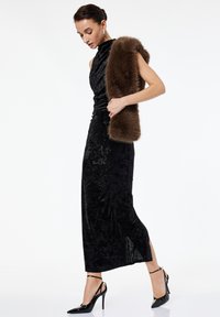 Svart sammetsklänning med hög halsringning och rynkad design, kombinerad med en brun pälsstola. Svarta lackskor med ankelsnören fulländar looken.