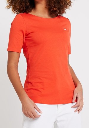 Kvinde iført en tætsiddende, klar orange kortærmet t-shirt og hvide bukser, stående med hænderne nær lommerne mod en ensfarvet baggrund.