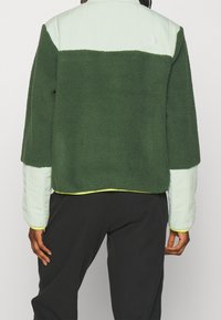 Zielona polarowa kurtka z jasnodołżonym pikowanym górnym odcinkiem, cechująca się zaokrąglonym krojem, mankietami i odblaskowym logo z tyłu.