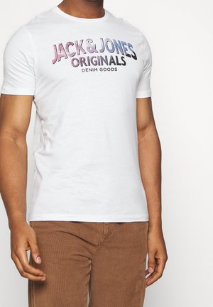 Mężczyzna w białej koszulce z tekstem "Jack & Jones Originals Denim Goods" i brązowych spodniach sztruksowych na białym tle.