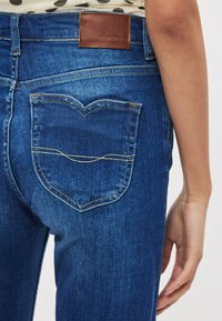 Kék farmernadrág, amely szív alakú hátsó zsebbel, barna bőr márkajelzéssel és látható varrási részletekkel rendelkezik.