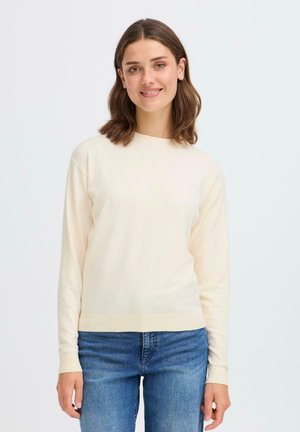 Kvinne med skulderlangt brunt hår iført en kremfarget langermet genser og blå jeans, smilende mot en enkel lys bakgrunn.